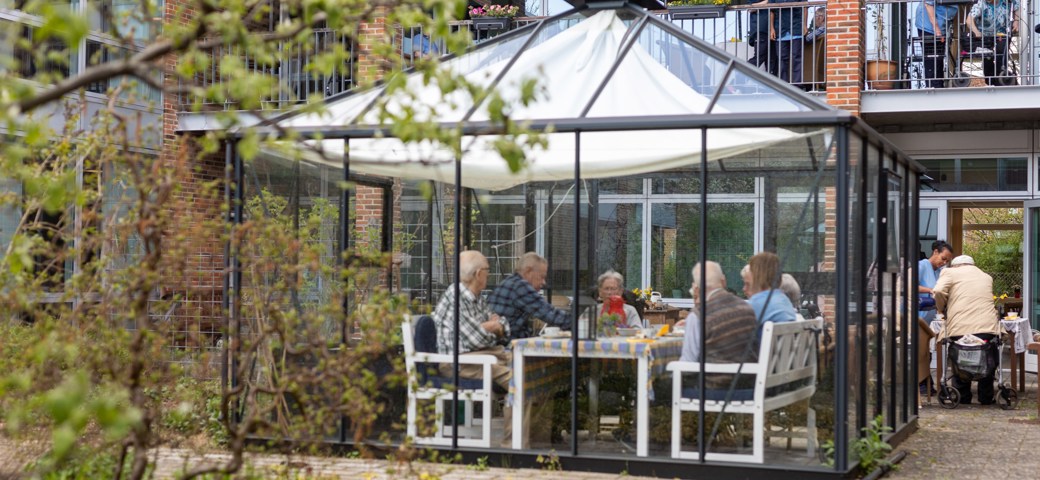 Ældre borgere nyder eftermiddagskaffen i drivhuset - samme billeder blot lidt længere afstand