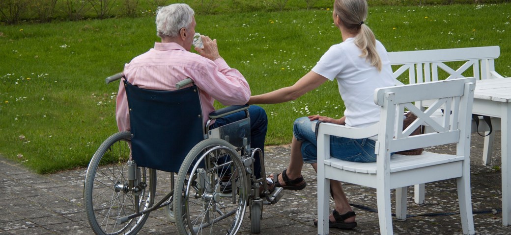 To kvinder sidder og taler sammen, en ung kvinde på en hvid bænk og en ældre kvinde i en rullestol